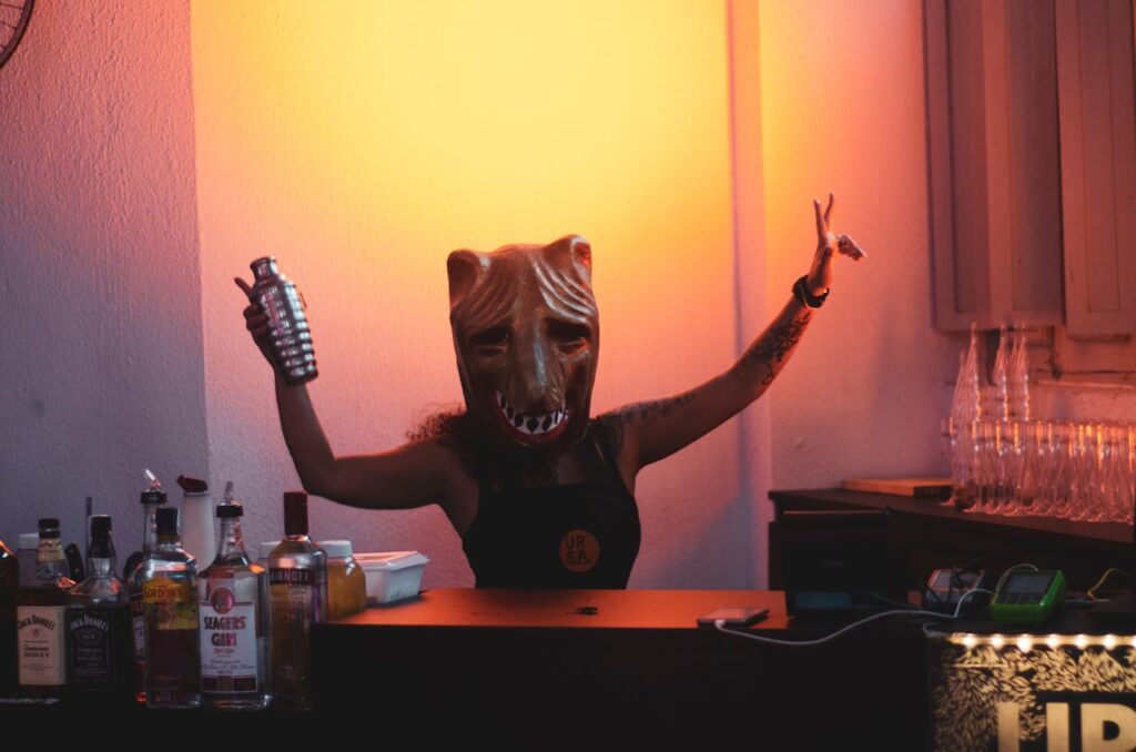 Person wearing an animal mask while bartending, surrounded by various bottles and creating a lively party atmosphere under warm orange lighting.