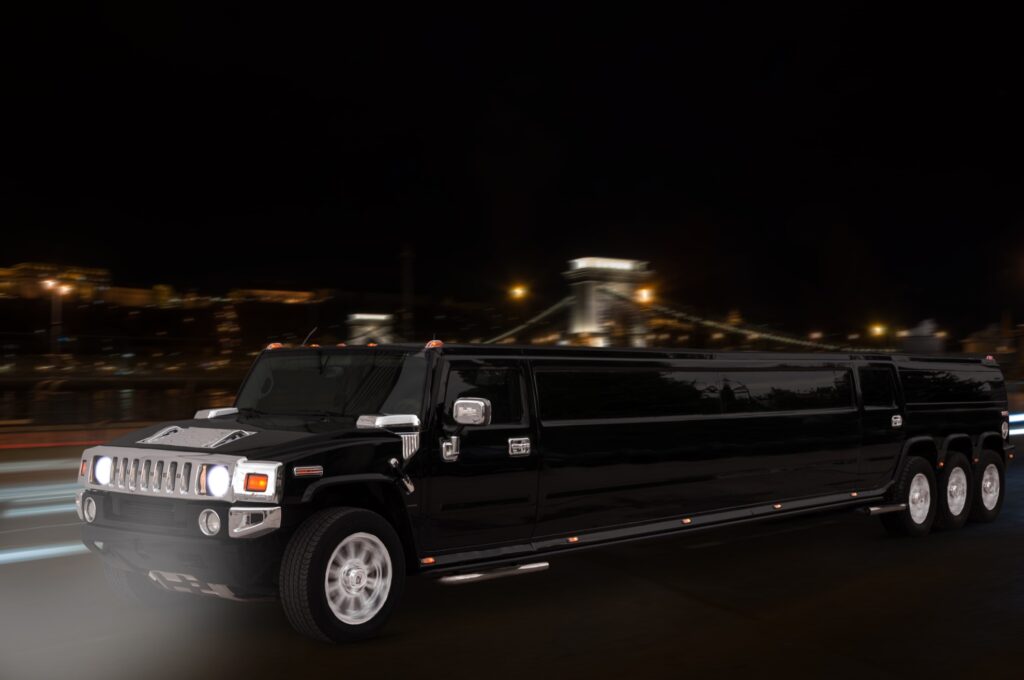 Black Hummer limousine driving through Budapest at night, with the illuminated Chain Bridge in the background, highlighting luxury and elegance.