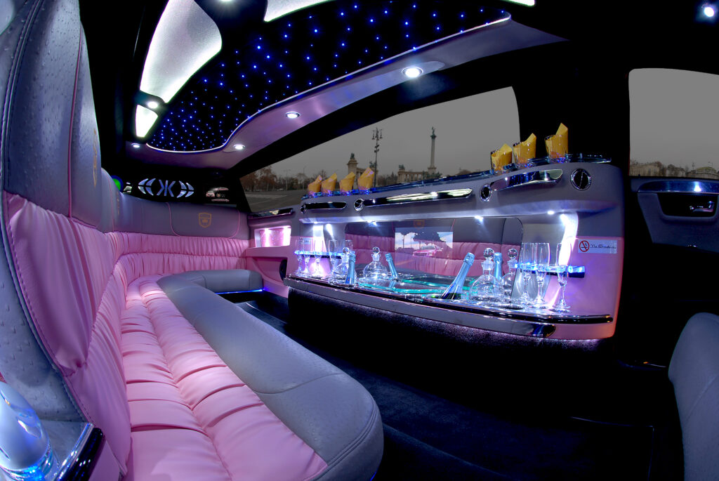 An elegant limousine interior featuring pink leather seating, ambient blue lighting on the ceiling, and a fully stocked bar with glassware, creating a luxurious and festive atmosphere.