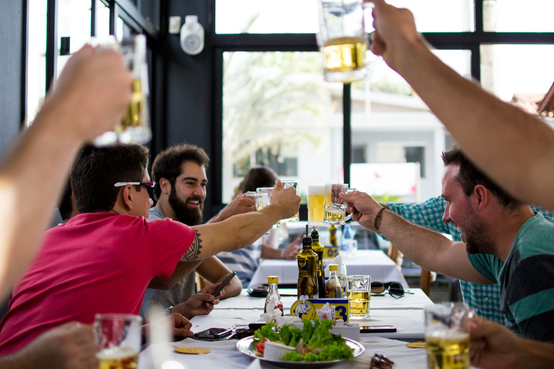 A group of friends raising their beer mugs in celebration at a lively gathering, enjoying good food and camaraderie in a bright, modern venue.