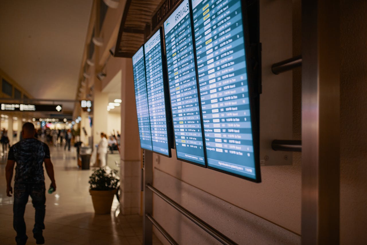 An airport arrivals board displaying flight information, with passengers walking in the background through a brightly lit terminal.