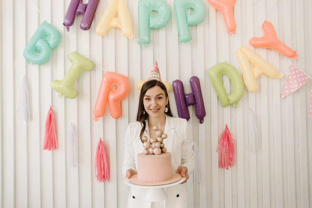 A woman celebrating her birthday party with colorful Happy Birthday balloons in the background, holding a decorated pink cake with a cheerful smile.