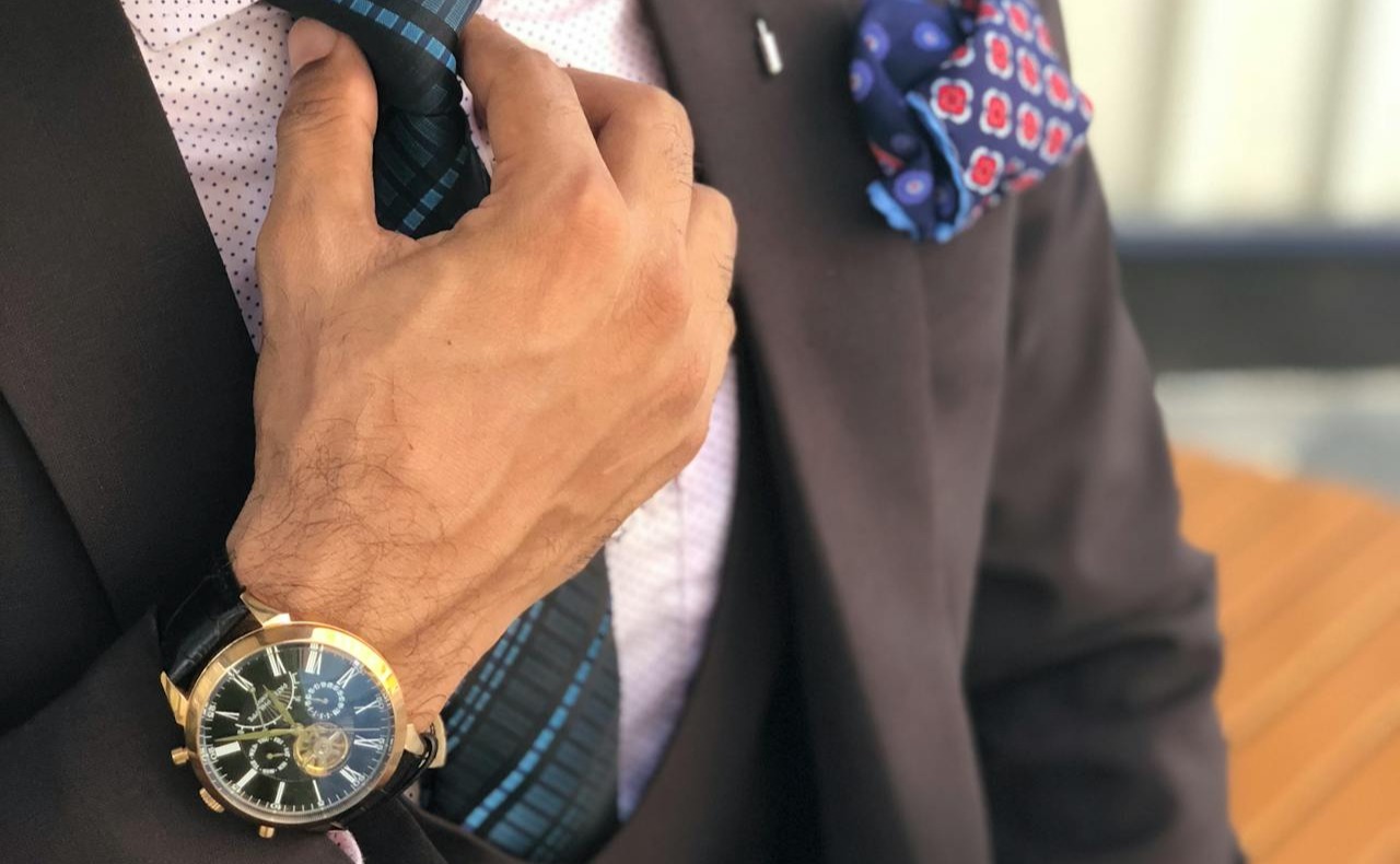 A close-up of a stylishly dressed man adjusting his tie, showcasing elegant details like a patterned pocket square and a luxury wristwatch, exuding sophistication and attention to detail.